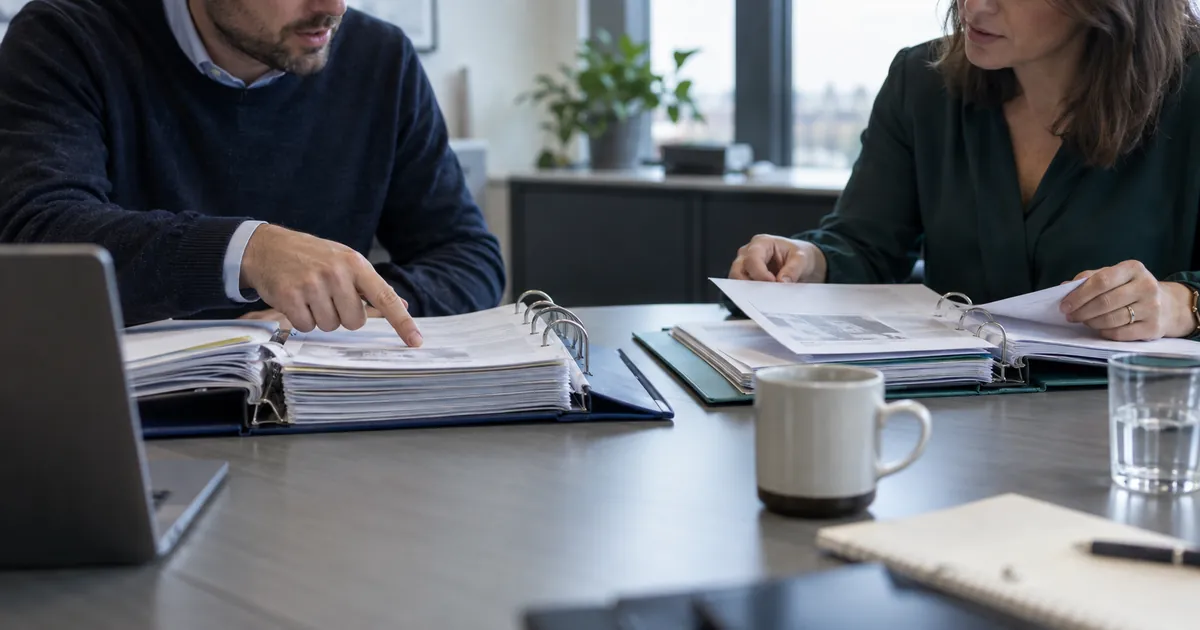 Two colleagues at a meeting table compare pages in two different procedure binders.